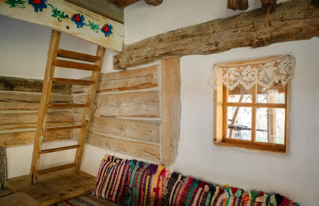 Interior de cameră tradițională din bârne de lemn, cu perne țesute colorate și fereastră cu perdea de dantelă