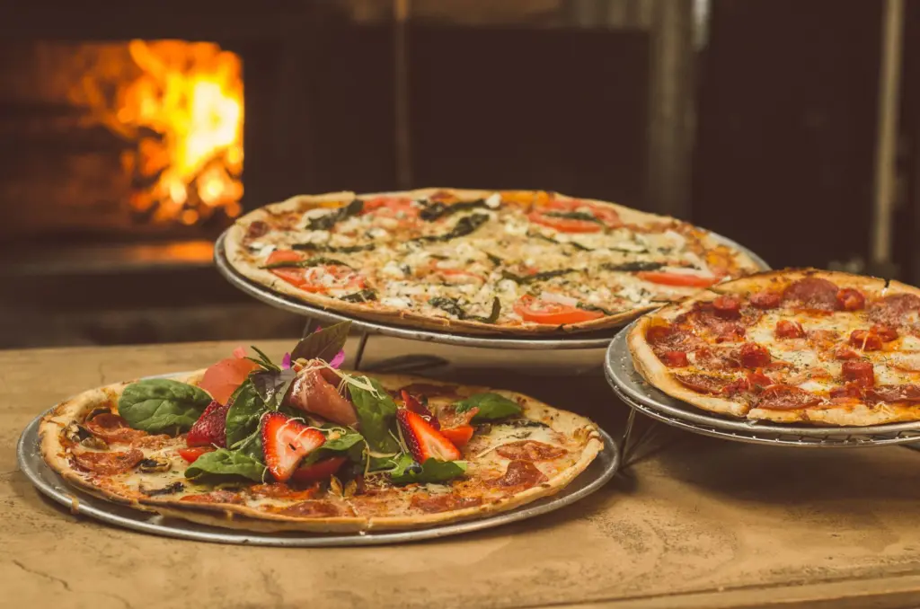 Acasă (v2) Trei pizza proaspăt coapte pe suporturi, în fața unui cuptor cu lemne aprins